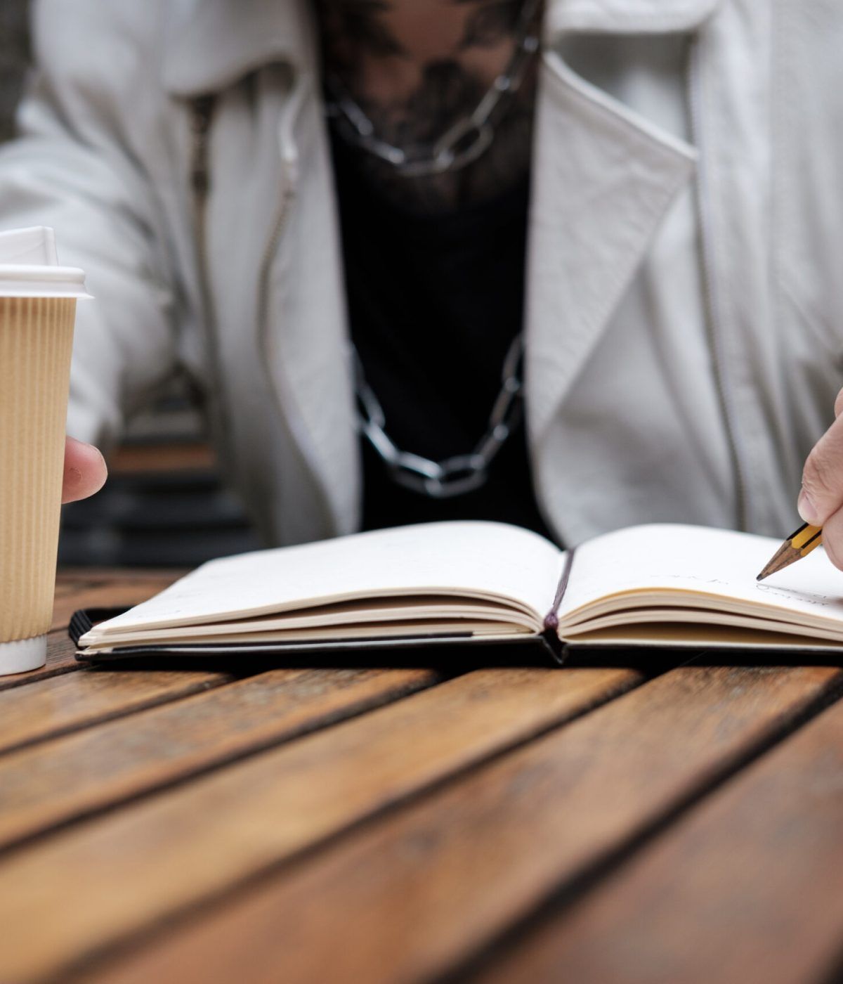 Concept portrait of unrecognizable tattoo writing on notepad. No technology involved, old fashioned habit concept. He is outside, on a table, holding a coffee.